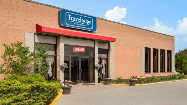 A Travelodge by Wyndham hotel with a brick facade, entrance marked by a red awning, and potted plants in front.