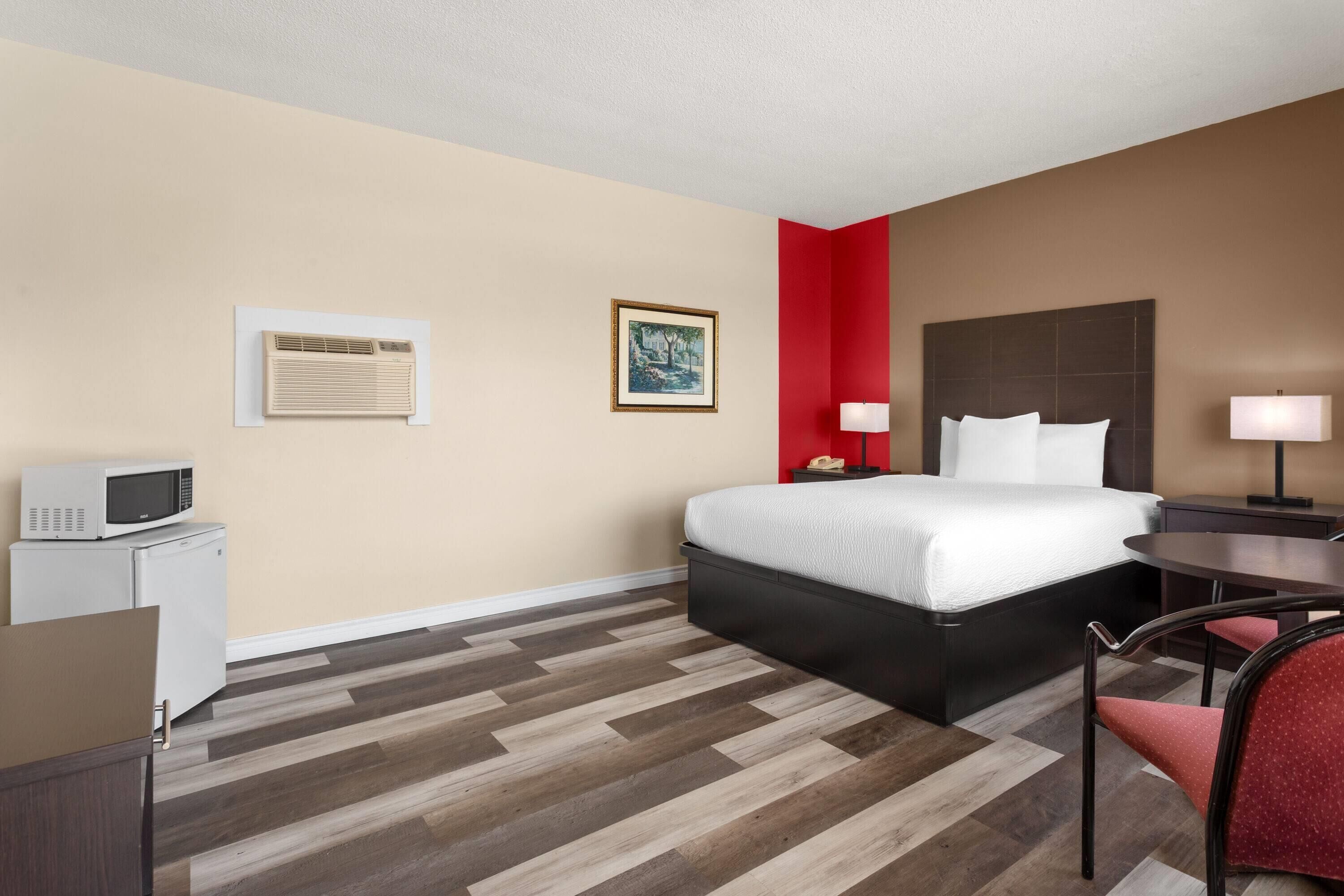 A hotel room with a bed, a desk with a microwave, a chair, a bedside table with a lamp, and a framed picture on the wall.