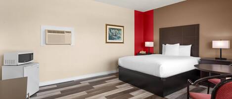 A hotel room with a bed, a desk with a microwave, a chair, a bedside table with a lamp, and a framed picture on the wall.