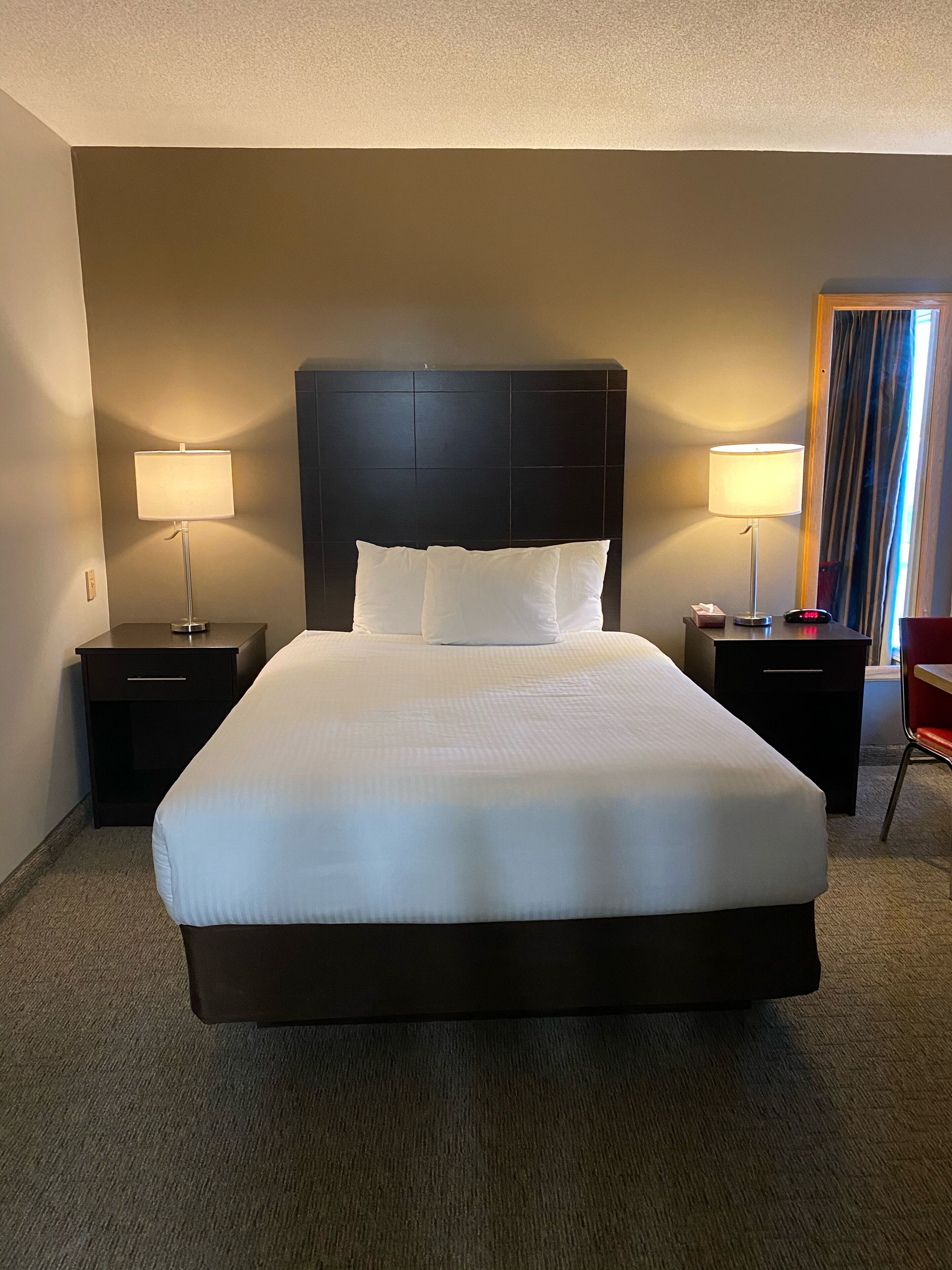 Une chambre d’hôtel avec un grand lit, deux tables de chevet avec des lampes, une tête de lit et une fenêtre avec des rideaux.