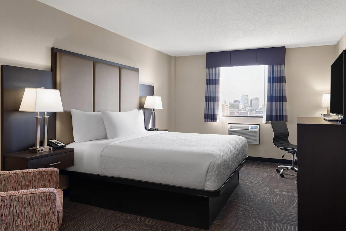 A hotel room with a large bed, two bedside tables with lamps, a desk with a chair, and a view of the city through the window.