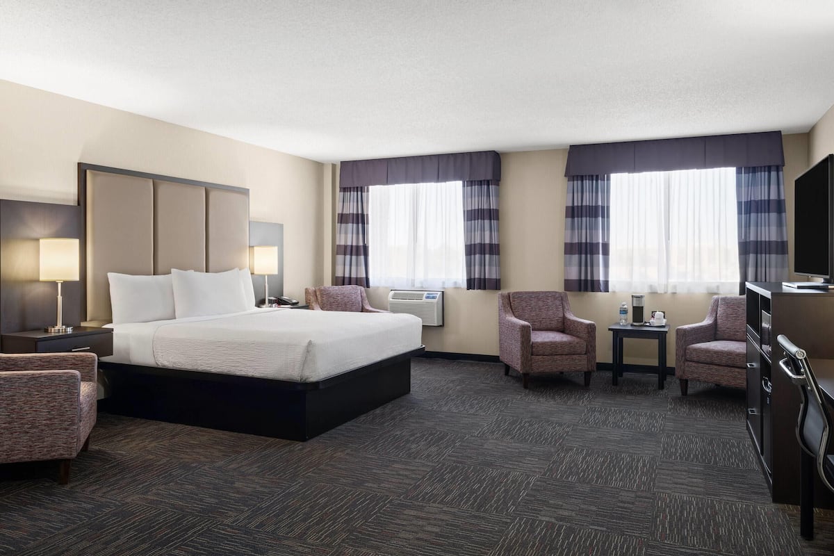 A hotel room with a large bed, two armchairs, a desk, a TV, and two windows with curtains.