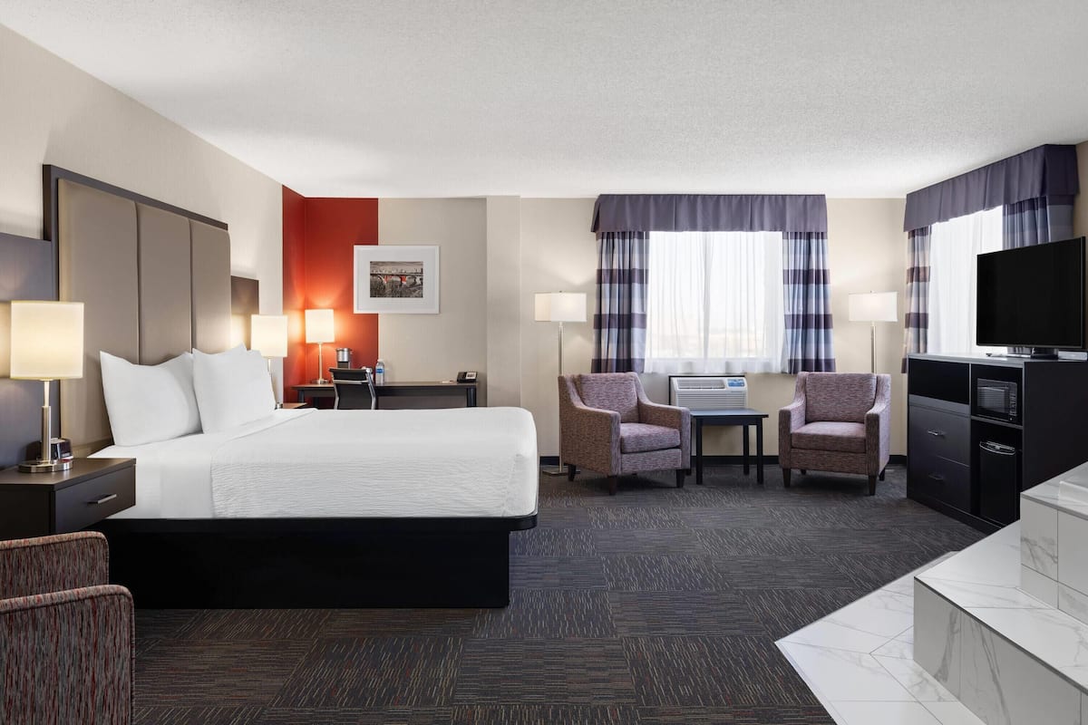 A hotel room with a large bed, two armchairs, a desk with a lamp, a television, and a minibar.