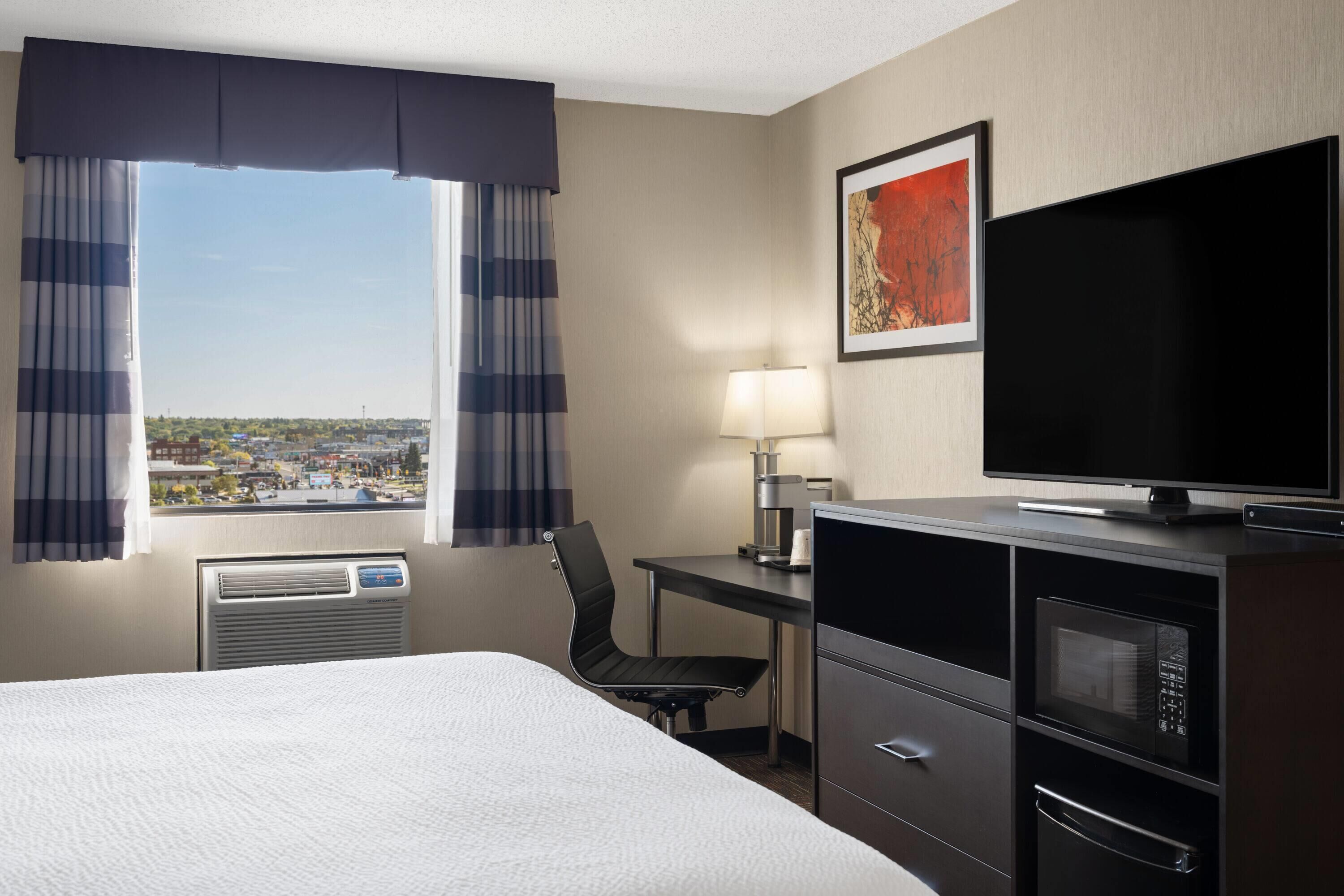 Habitación de hotel con cama, escritorio con lámpara, televisión, microondas y vista a la ciudad.