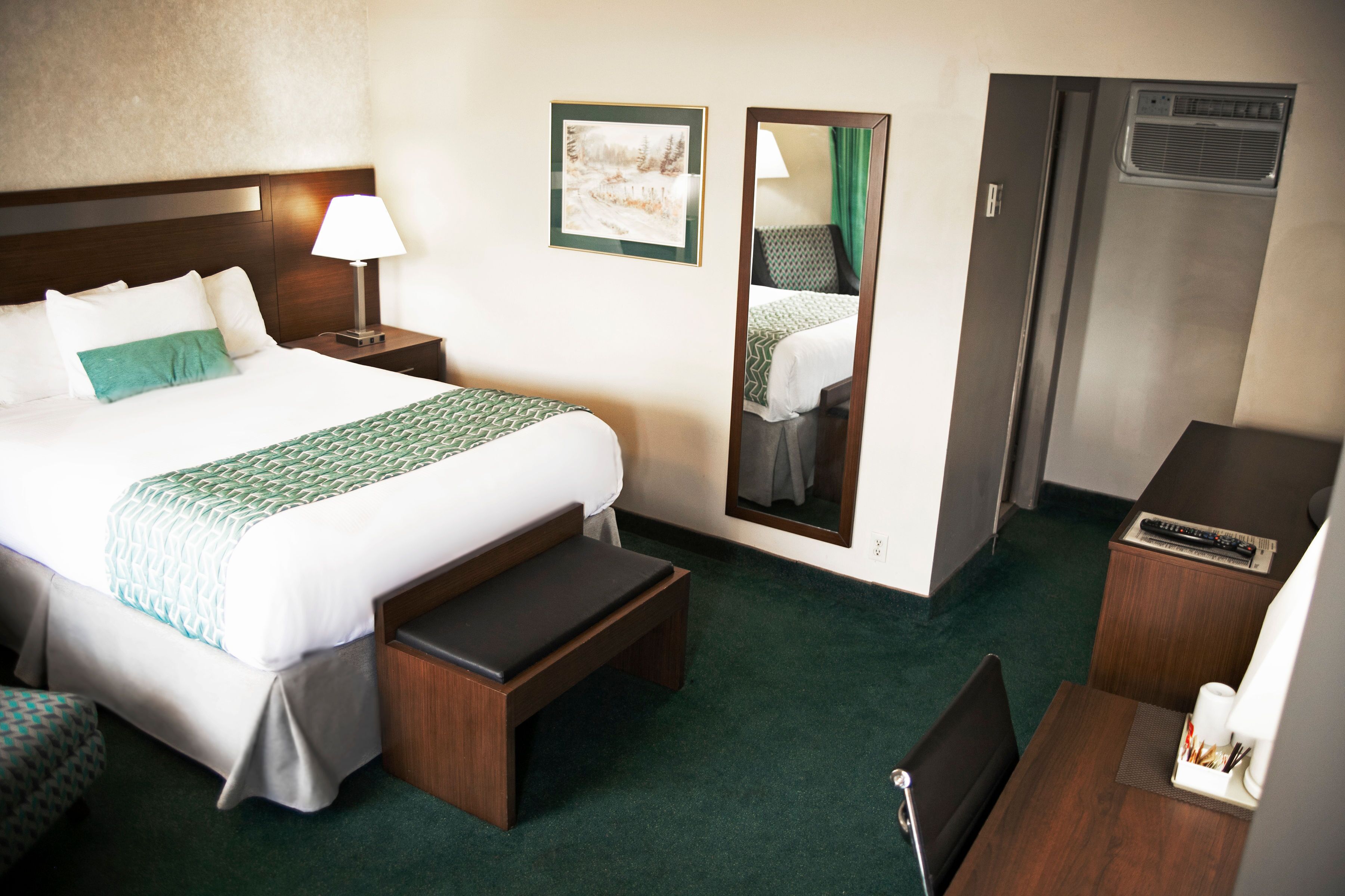 Quarto de hotel com uma cama grande, uma mesa de cabeceira com abajur, um espelho, uma escrivaninha com cadeira e um pequeno banco.