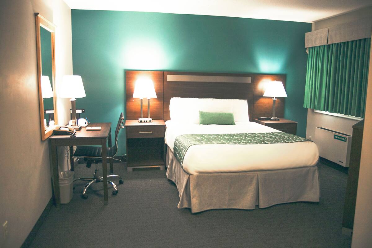 Una habitación de hotel con una cama, dos mesitas de noche con lámparas, un escritorio con una silla, un espejo y un cubo de basura.