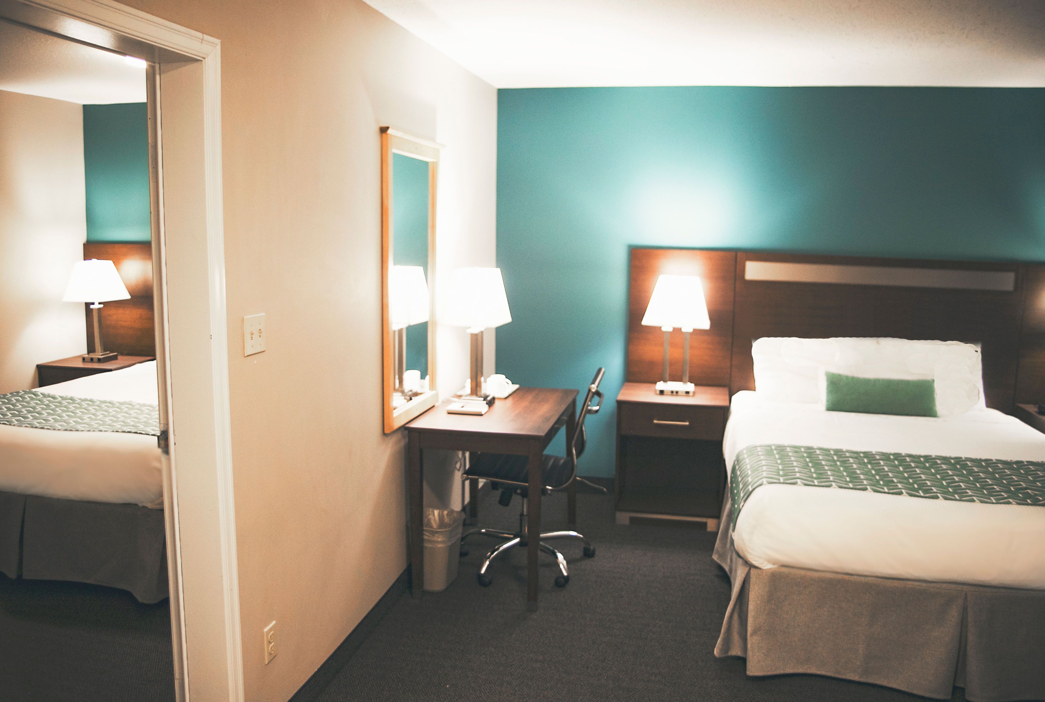 A hotel room with a bed, a desk, and a chair. There is a mirror above the desk and a lamp on the desk. The room has a blue wall and a wooden headboard on the bed.