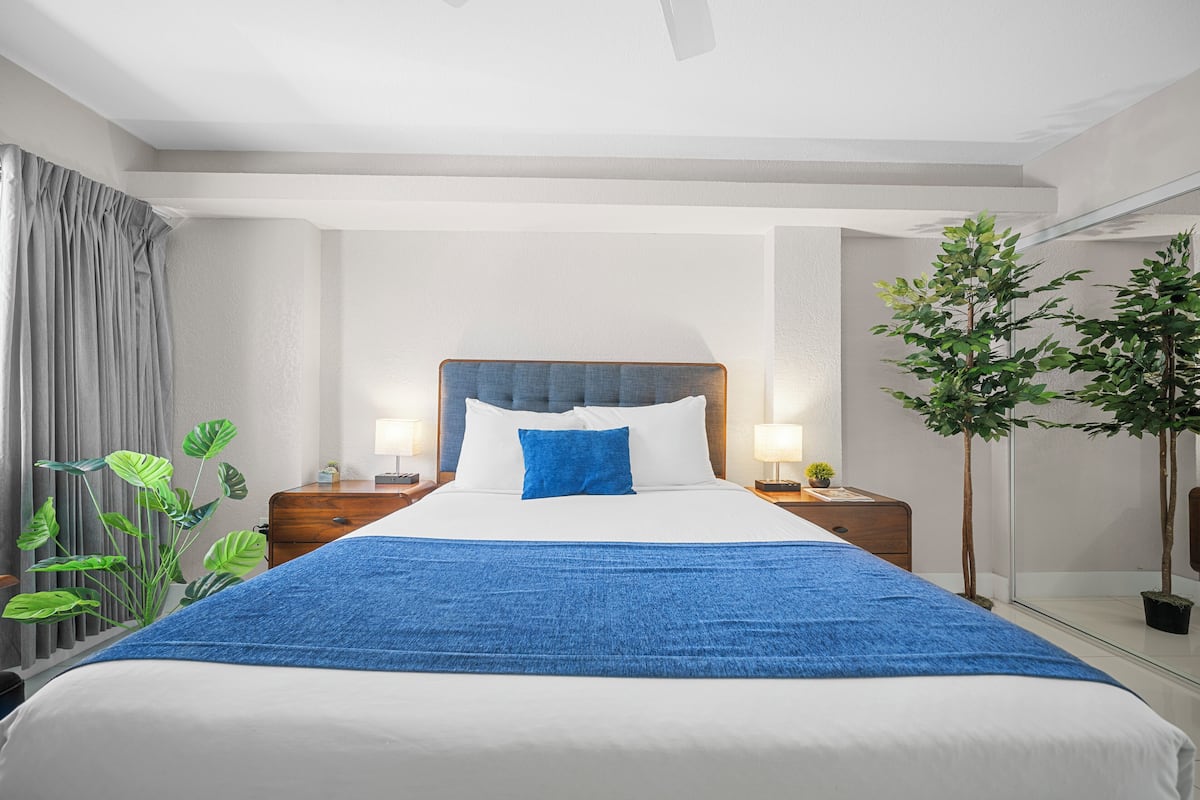 A hotel room with a large bed, a wooden nightstand, a lamp, a potted plant, and a mirror.
