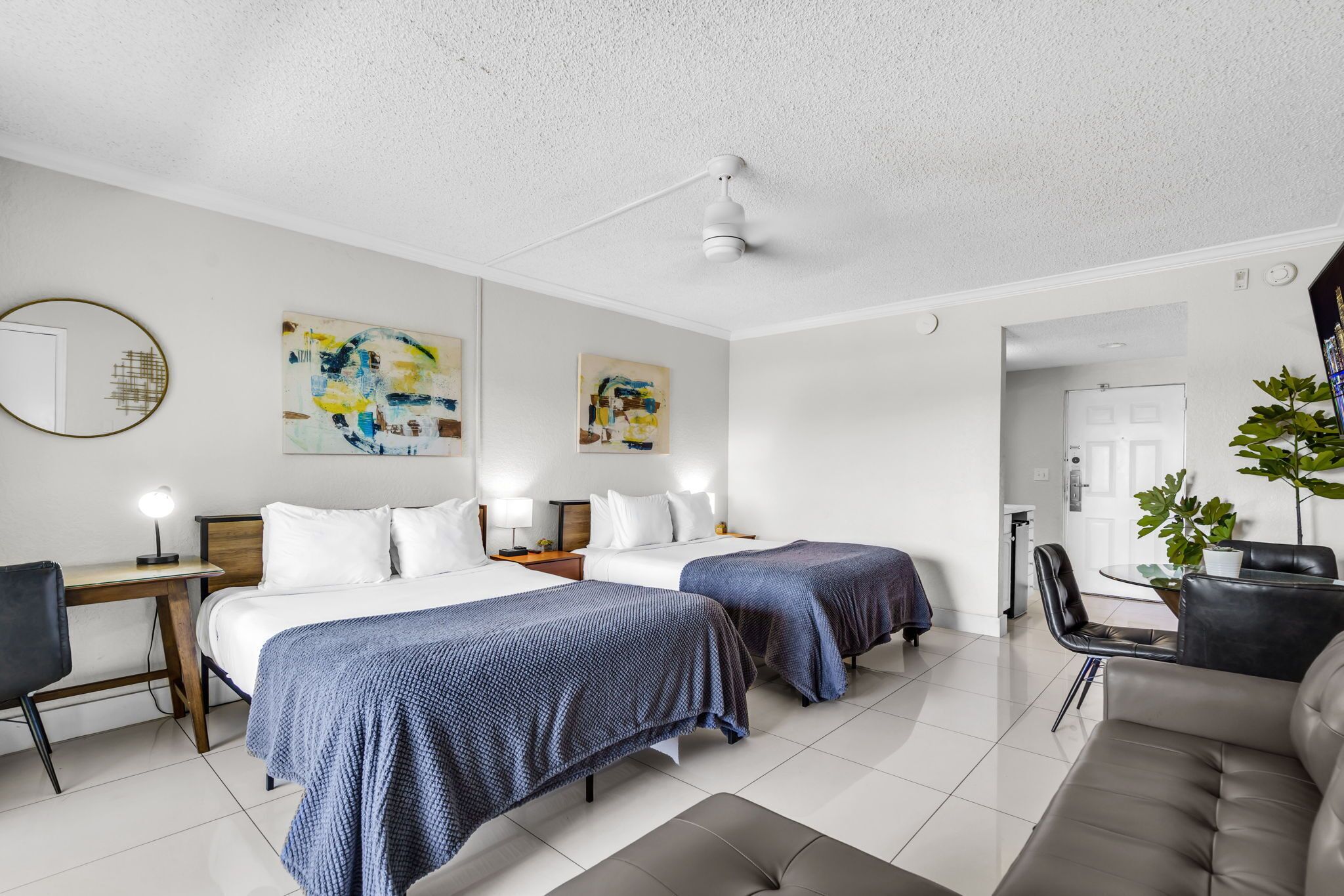 A modern hotel room with two beds, a desk, a chair, a sofa, and a dining table.
