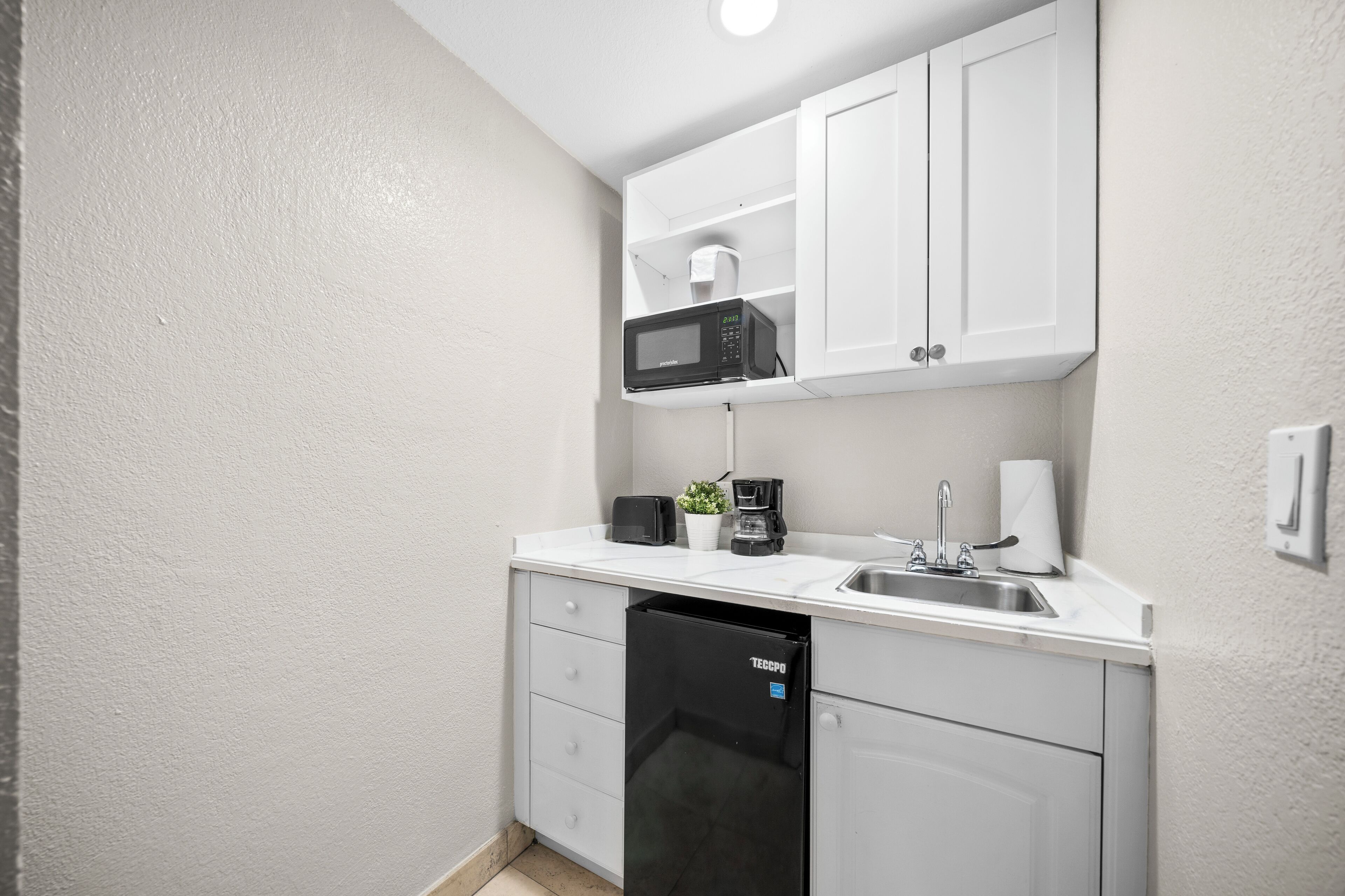 A compact kitchenette with white cabinets, a microwave, a coffee maker, a sink, and a dishwasher.
