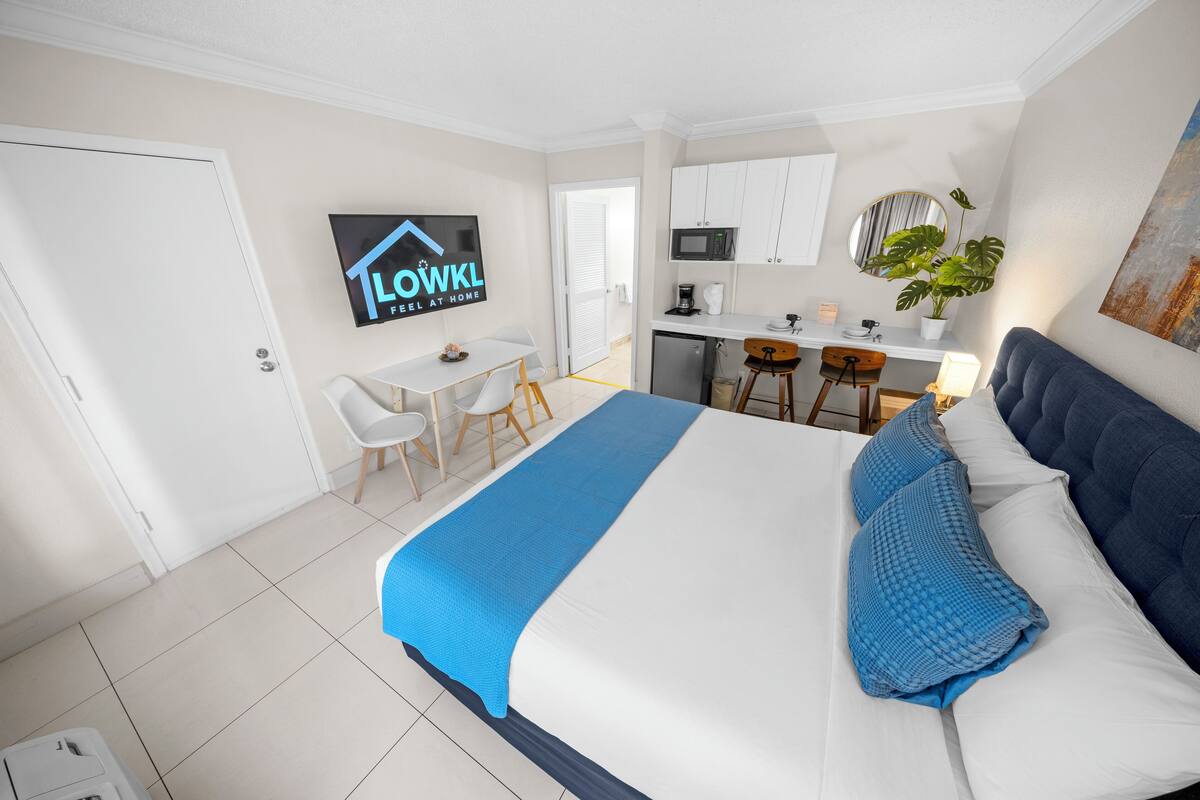A hotel room with a bed, a desk, a chair, a microwave, and a potted plant.