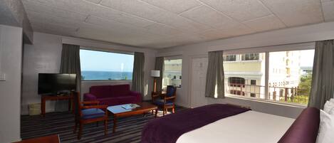 A hotel room with a large bed, a TV, a sofa, and a view of the sea.