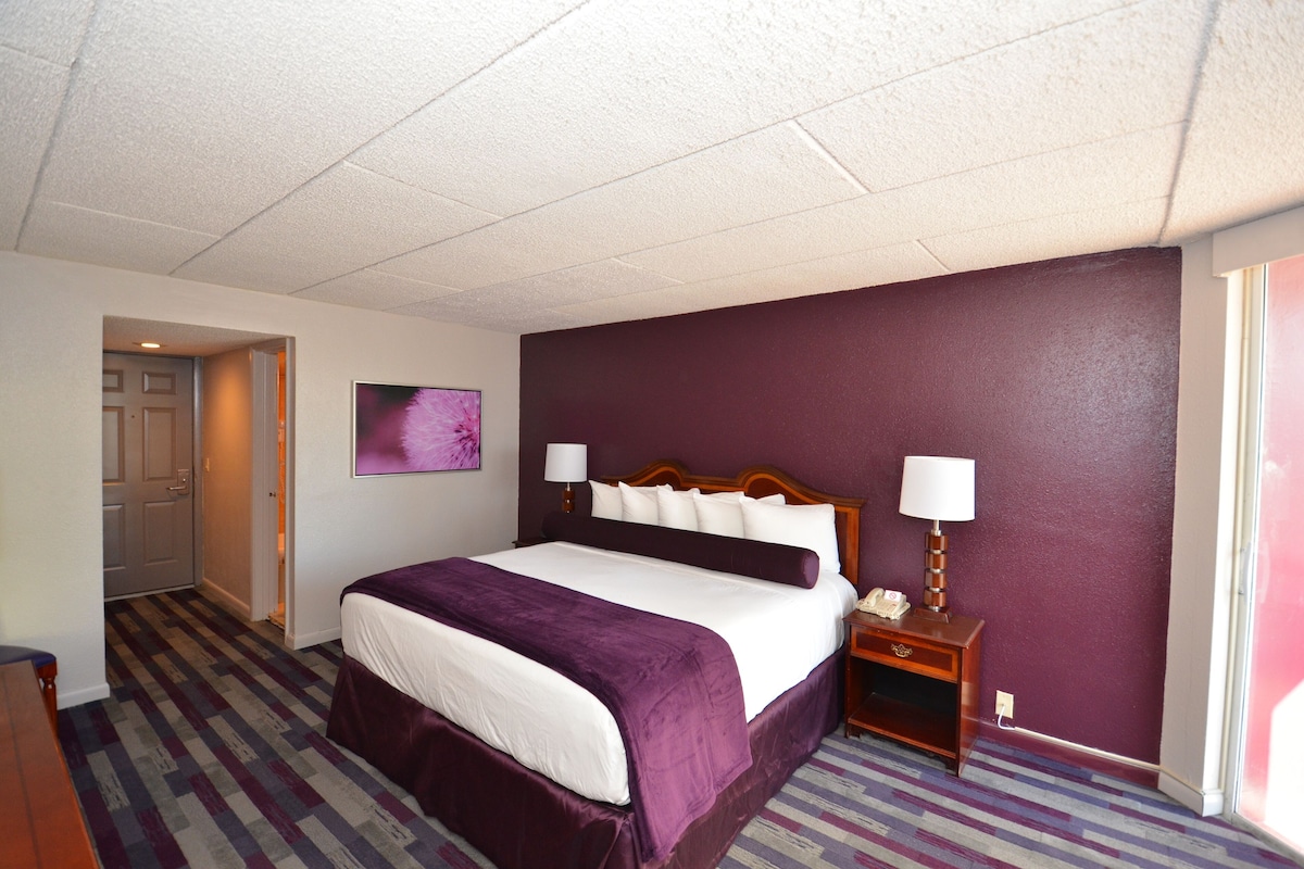 A hotel room with a large bed, a nightstand, a door, and a wall with a purple accent.