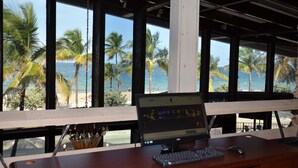 A laptop on a desk with a view of a beach and palm trees through large windows.