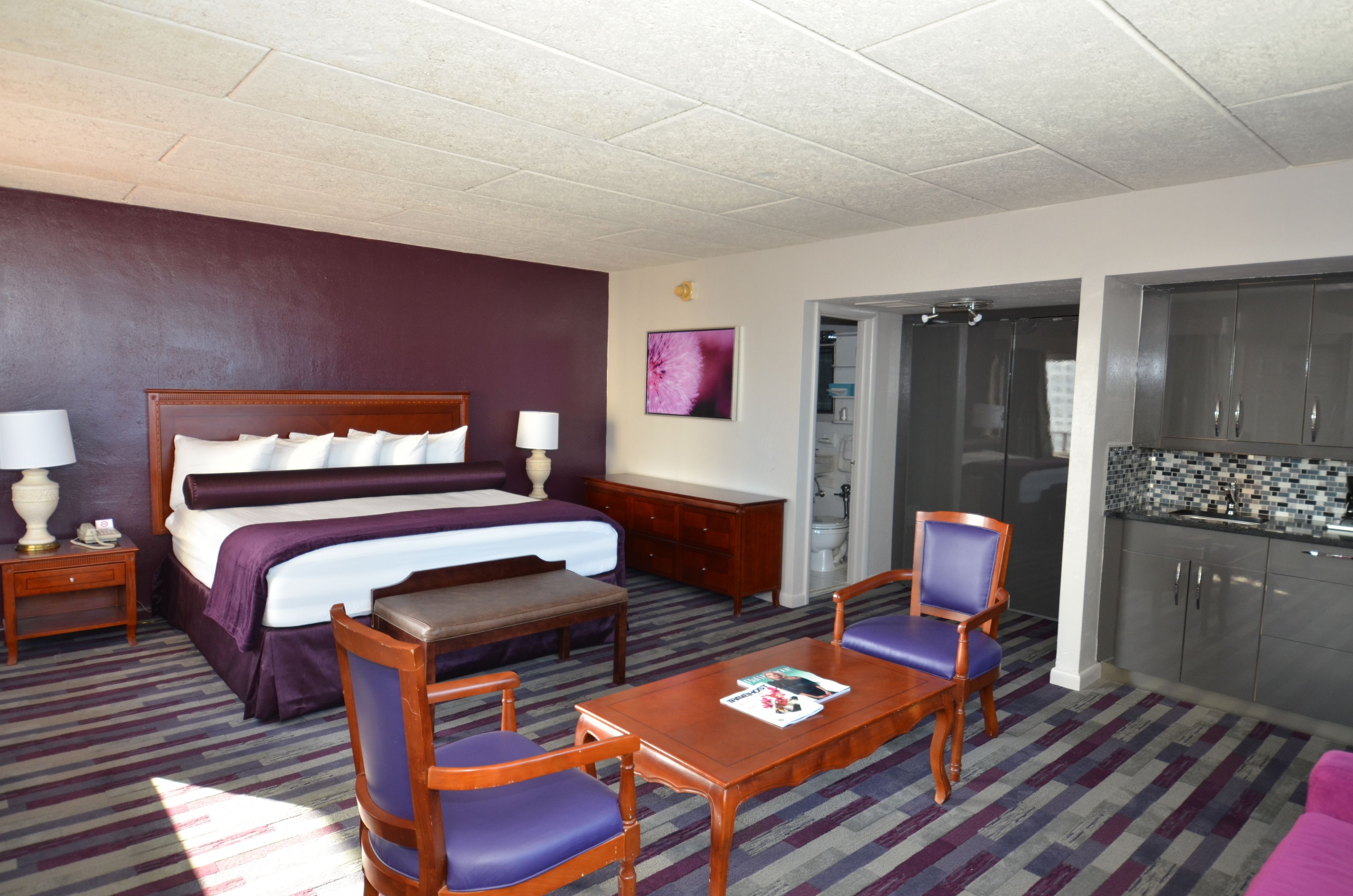 Une chambre d’hôtel comprenant un lit, des tables de chevet, un bureau, des chaises, une petite table, une salle de bain avec douche et une kitchenette.