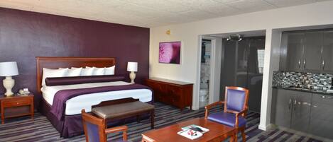 Une chambre d’hôtel comprenant un lit, des tables de chevet, un bureau, des chaises, une petite table, une salle de bain avec douche et une kitchenette.