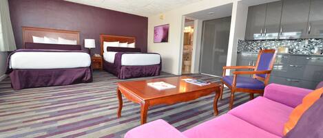 A hotel room with a bed, two bedside tables, a coffee table, a sofa, a chair, and a kitchenette.