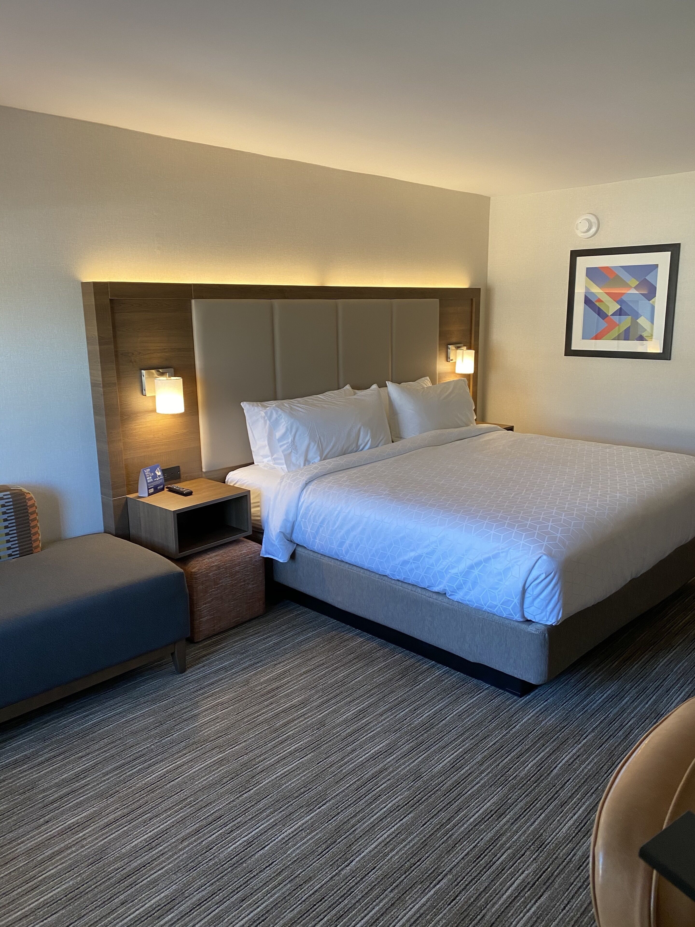 A hotel room with a large bed, a bedside table, a nightstand, a chair, and a wall-mounted lamp.