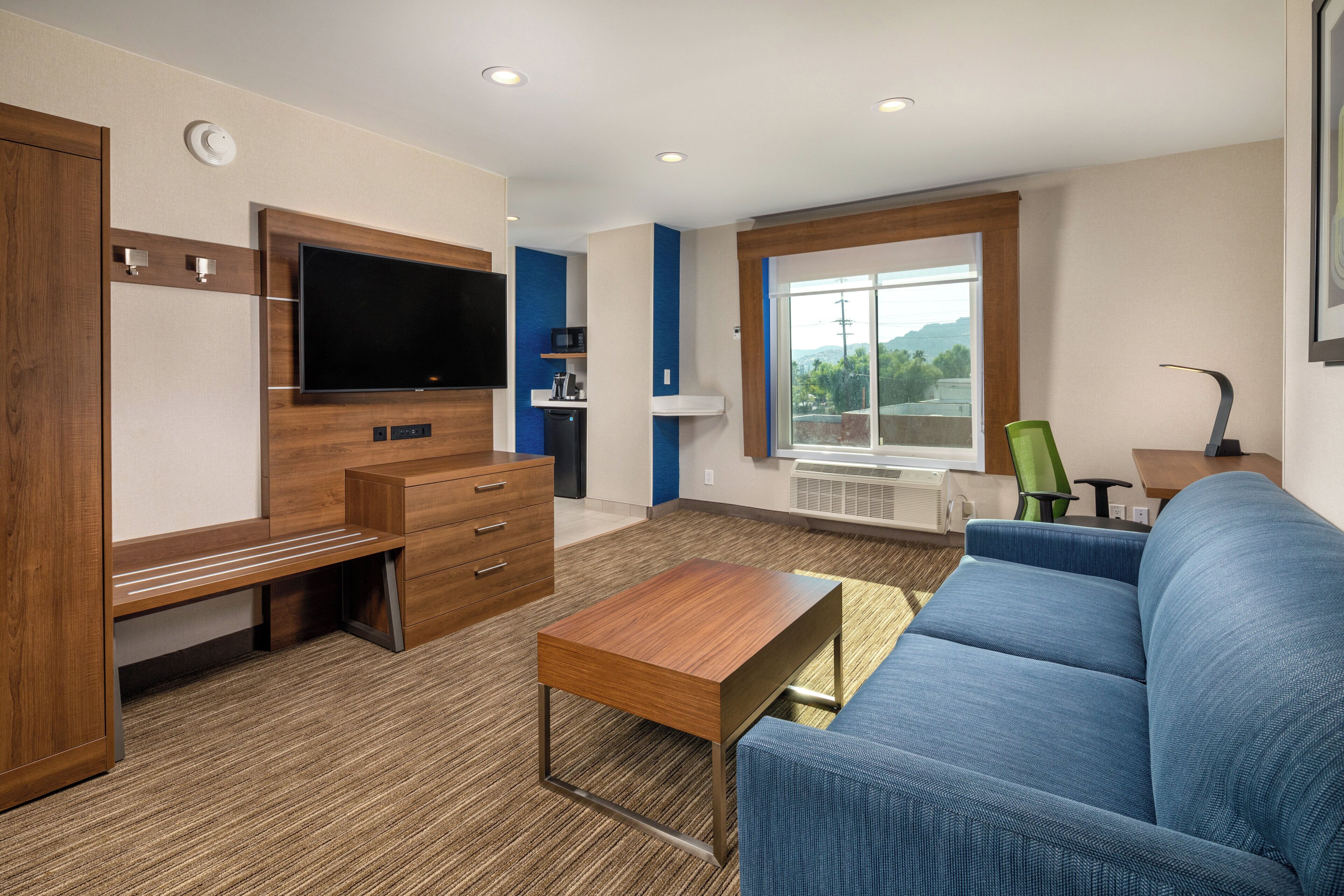 Una camera d'albergo moderna con una TV a schermo piatto, mobili in legno, un divano blu e un'ampia finestra con vista sull'esterno.