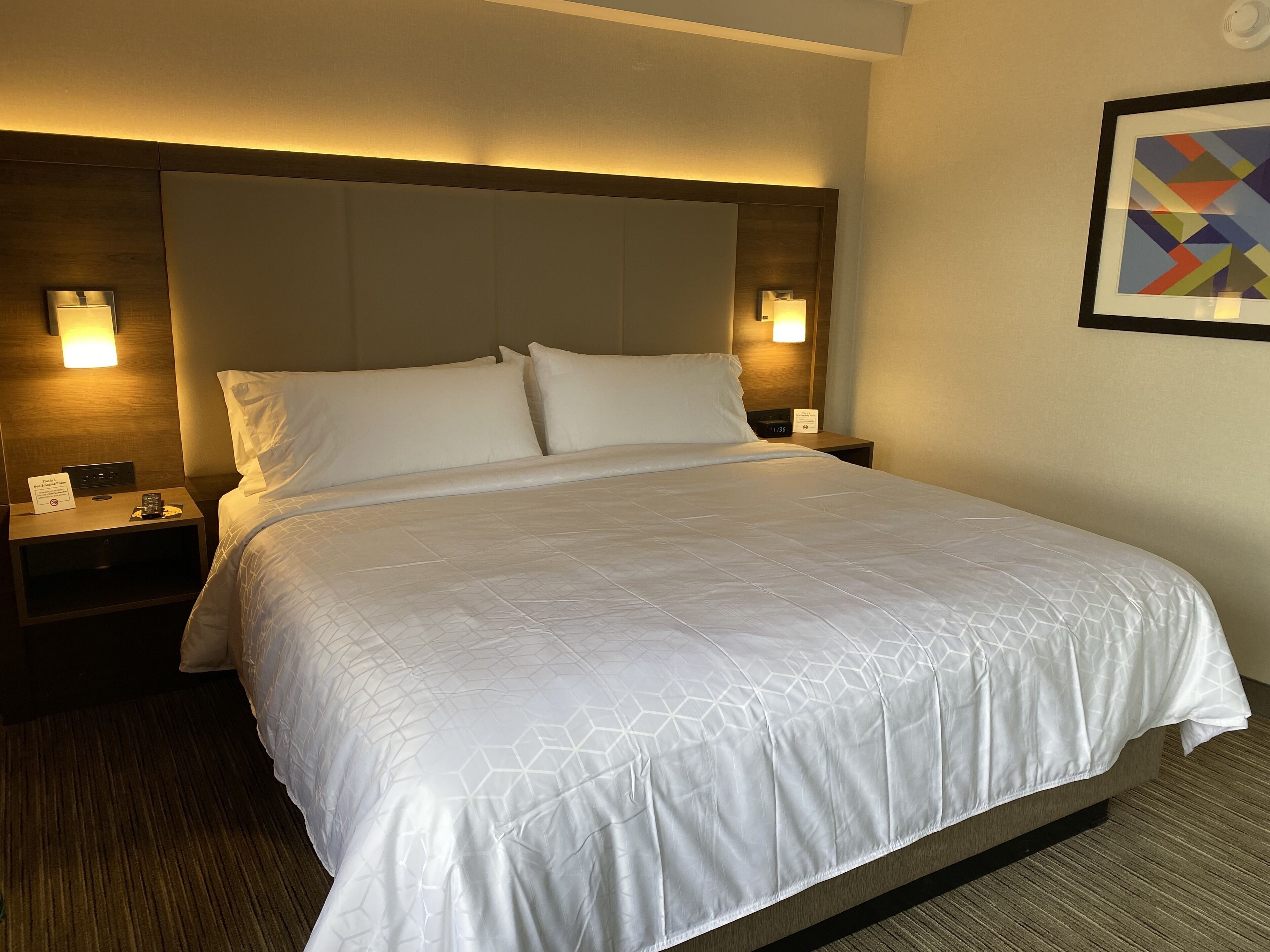 Ein Hotelzimmer mit einem großen Bett, zwei Nachttischen, einer Kommode, einer Wanduhr und einem gerahmten abstrakten Kunstwerk.