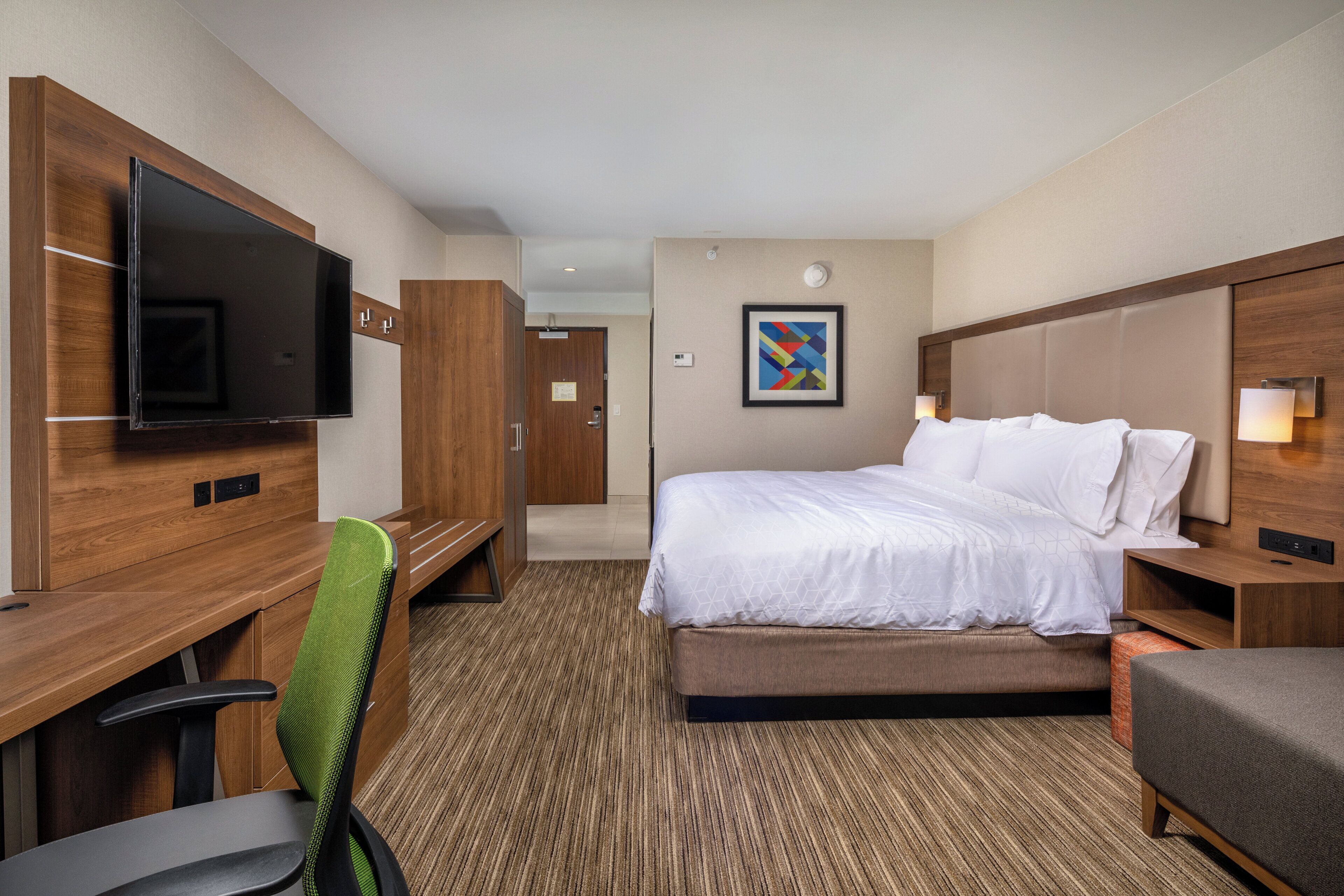 A hotel room with a bed, a desk with a flat-screen TV, a chair, and a bedside table.