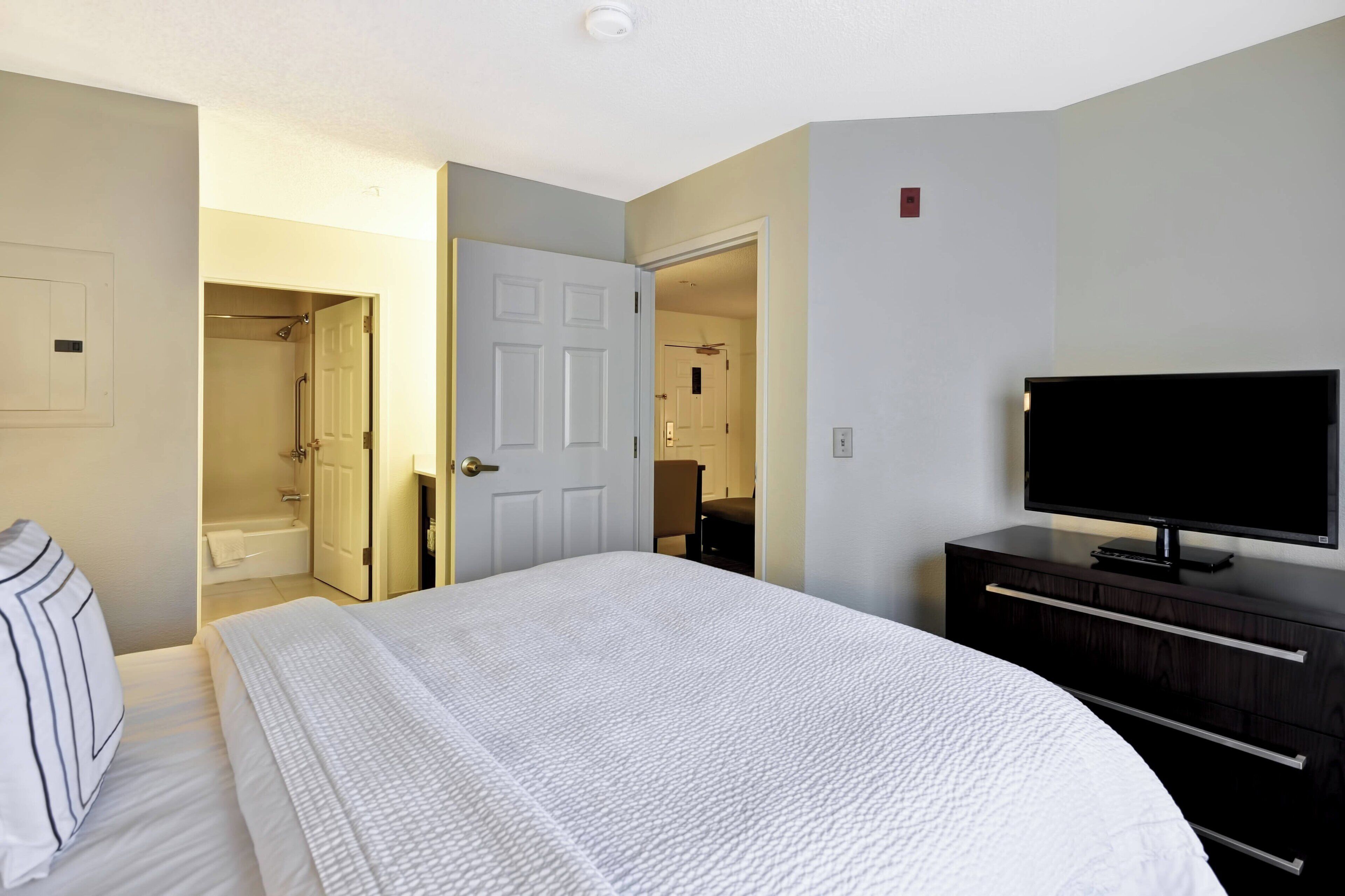 A hotel room with a large bed, a TV, a dresser, and a bathroom visible through an open door.