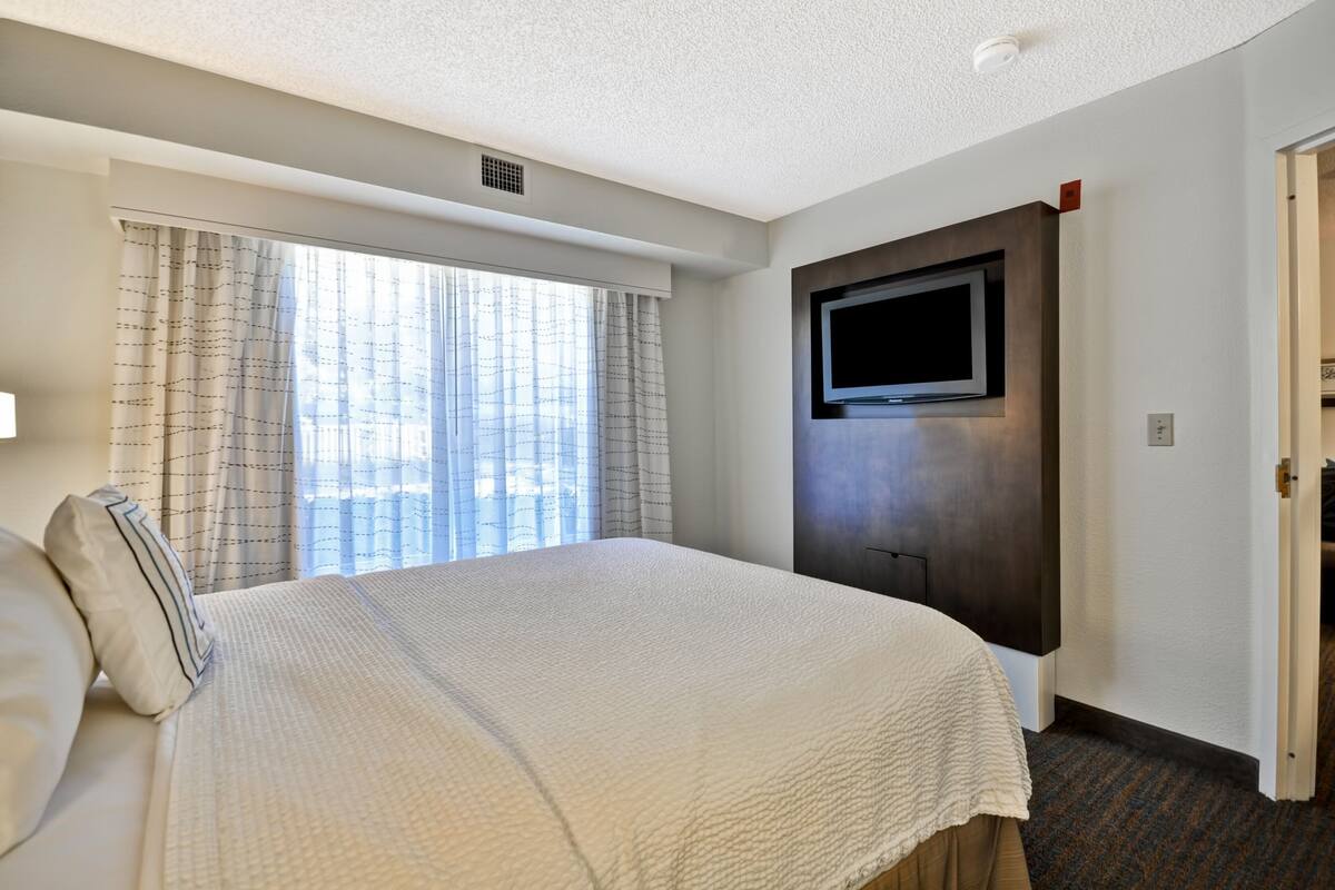 A hotel room with a large bed, a flat-screen TV mounted on the wall, and a window with curtains.