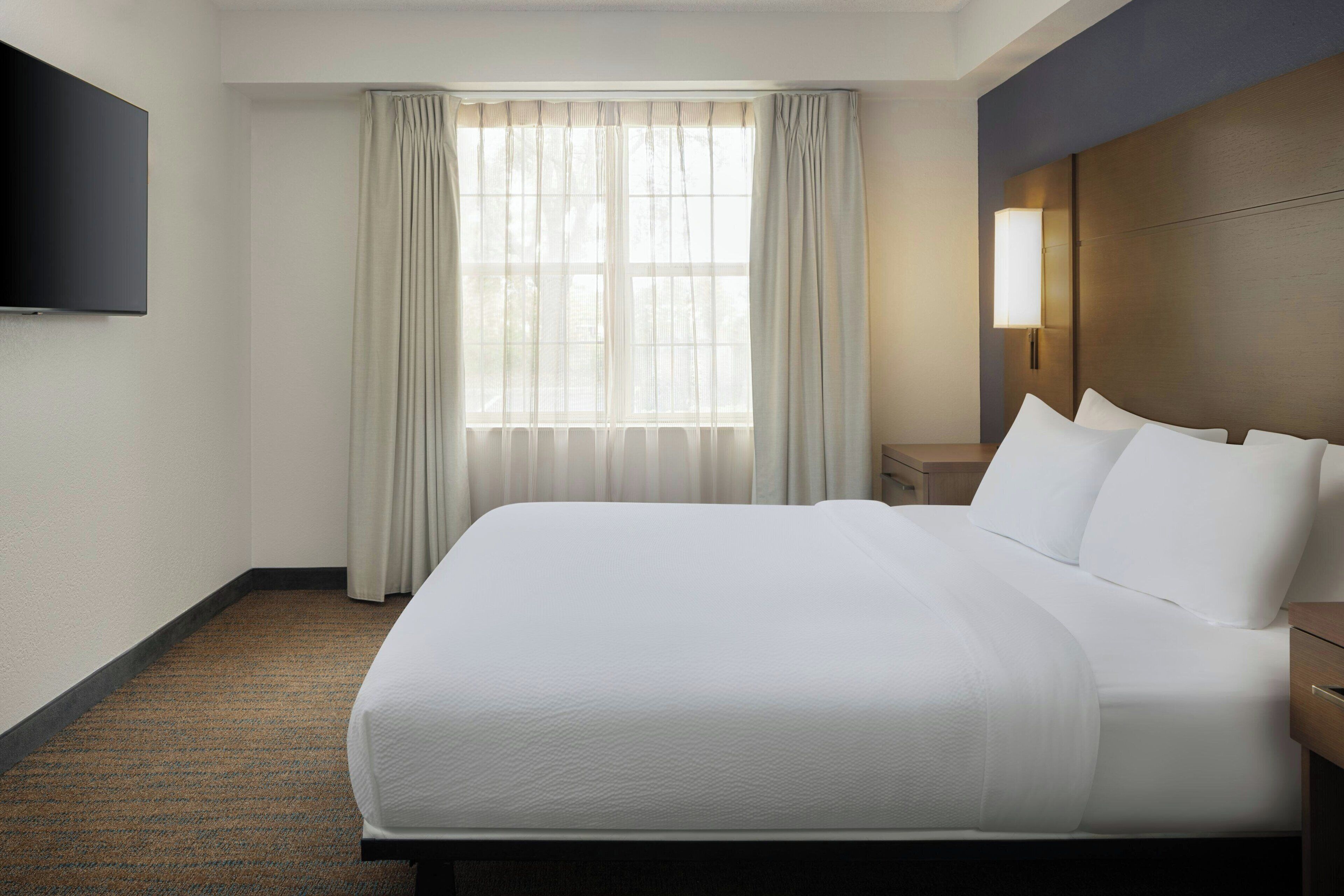 Ein Hotelzimmer mit einem großen Bett, einem flachbildfernseher an der Wand, einem Nachttisch und einem Fenster mit Vorhängen.