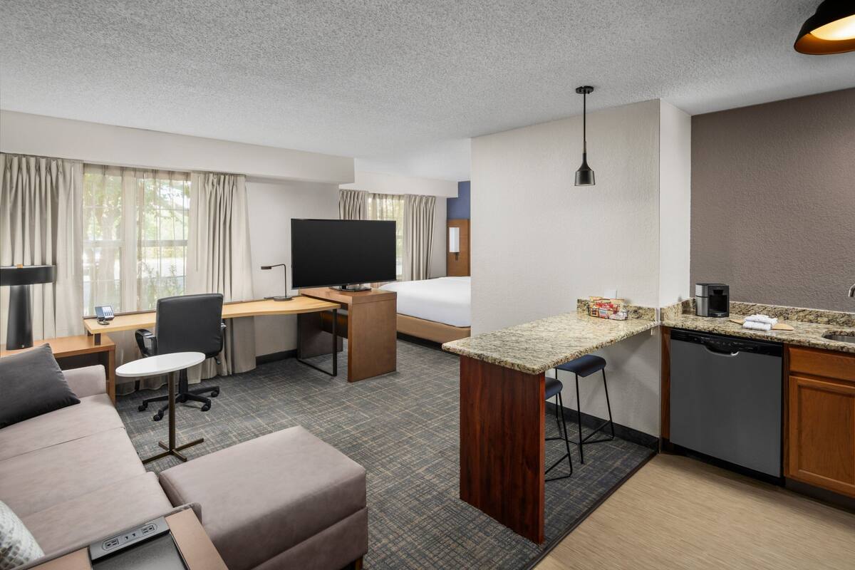 A hotel room with a sofa, a desk, a television, a kitchen area with a coffee maker, and a bed.