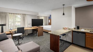 A hotel room with a sofa, a desk, a television, a kitchen area with a coffee maker, and a bed.