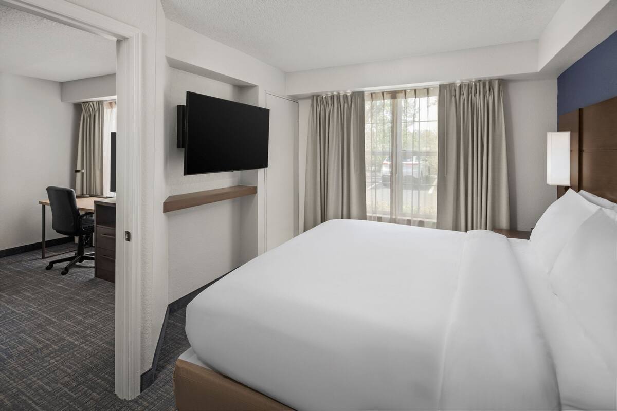 A hotel room with a large bed, a desk with a chair, a flat-screen TV mounted on the wall, and a window with curtains.