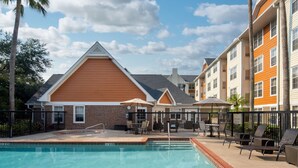 A pool area with lounge chairs and umbrellas, surrounded by a fence, and adjacent to multi-story buildings with a brick facade. - Residence Inn by Marriott Orlando East/UCF Area (Orlando)