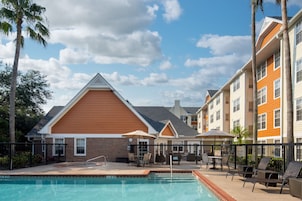 A pool area with lounge chairs and umbrellas, surrounded by a fence, and adjacent to multi-story buildings with a brick facade.
