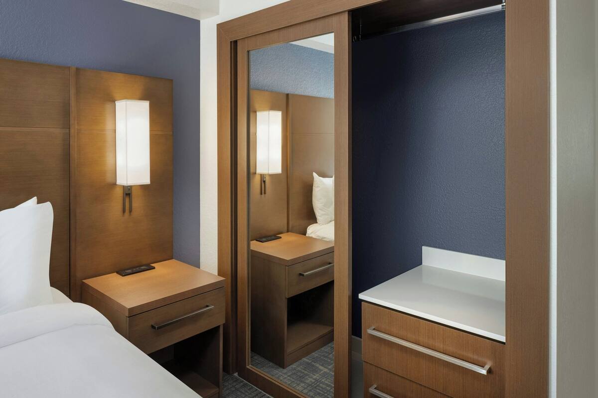 A hotel room with a bed, bedside table, nightstand, mirror, and a bathroom with a sink and countertop.