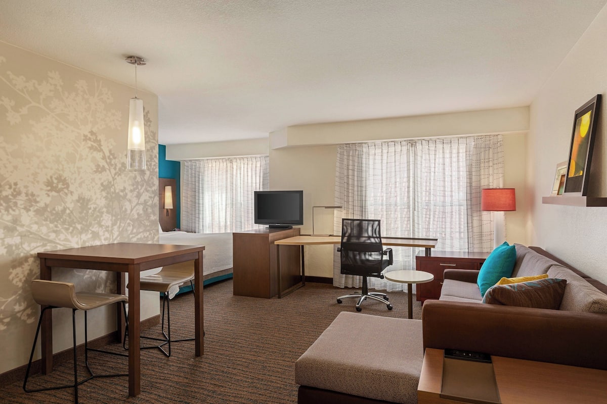 A hotel room with a bed, a desk with a computer, a TV, a sofa, and a small table.