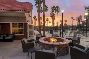 An outdoor patio with a stone fire pit surrounded by wicker furniture, a swimming pool in the background, and palm trees. - Residence Inn by Marriott Phoenix Chandler Fashion Center (Chandler)