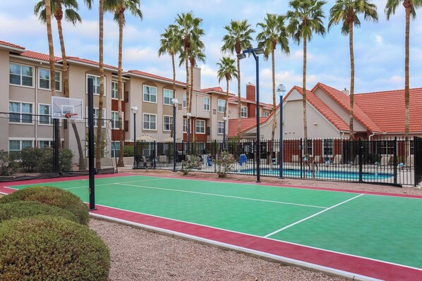 A tennis court with a basketball hoop, surrounded by palm trees and a pool.