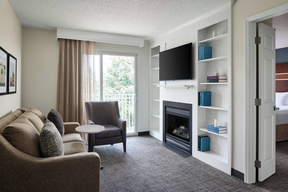 Hotelkamer met een bank, fauteuil, flatscreen-televisie, boekenkast, open haard en een deur naar een andere kamer.