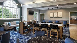 A spacious dining area with a large table, blue chairs, and a sofa. There is a TV mounted on the wall and a window providing a view of the outdoors. - Residence Inn By Marriott Raleigh Crabtree (Raleigh)