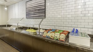 Un espace d’accueil d’hôtel comprenant un comptoir de buffet, une grande œuvre d’art encadrée au mur, et une variété de plats et de boissons proposés.