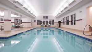 A large indoor swimming pool with clear blue water, two lounge chairs, and a staircase leading to an upper level.