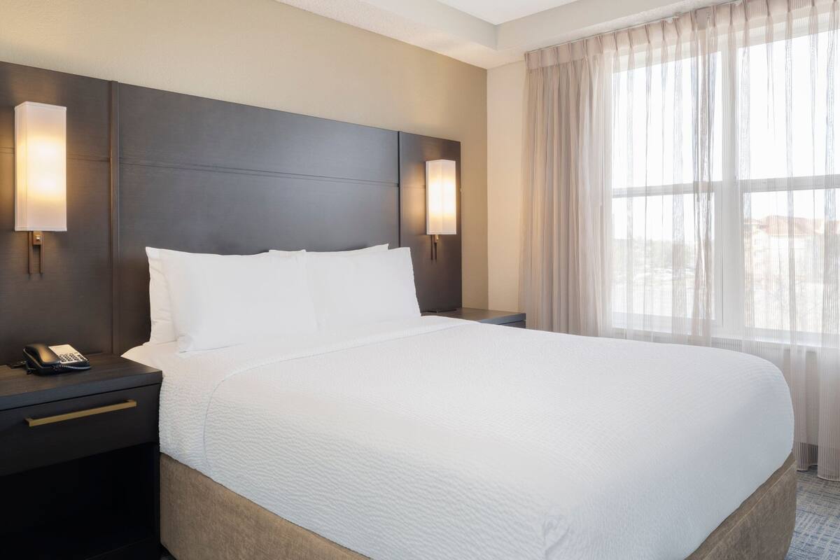 Quarto de hotel com uma cama grande, duas luminárias de cabeceira, mesa de apoio e uma janela com cortinas leves.