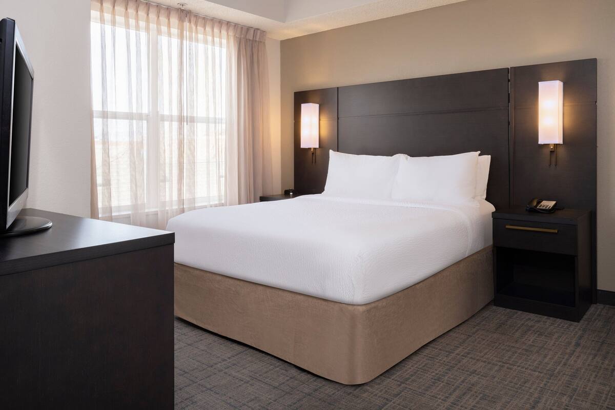 Quarto de hotel com uma cama grande, duas mesas de cabeceira, uma televisão e uma janela com cortinas leves.