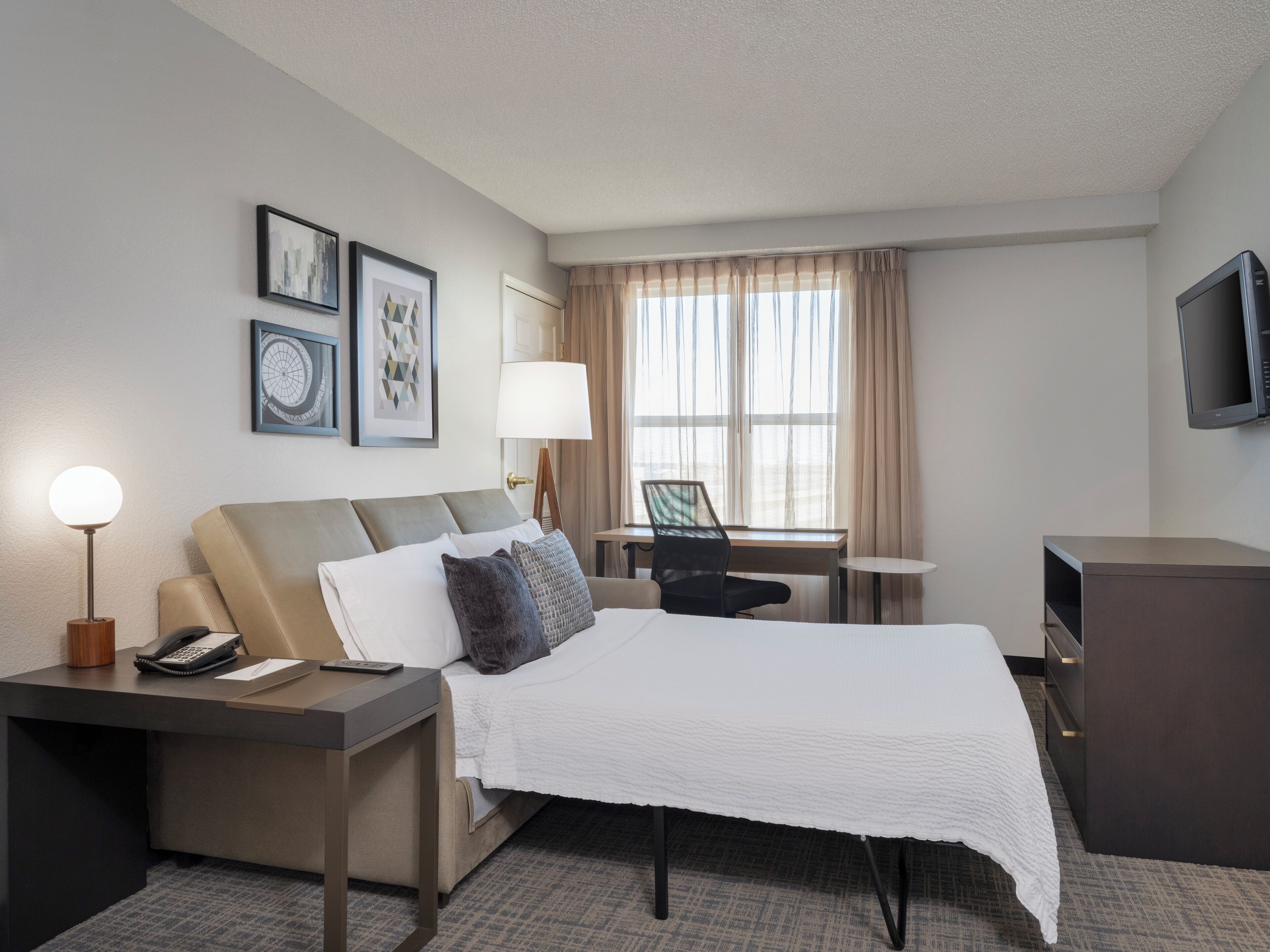 A hotel room with a bed, desk, chair, lamp, TV, and artwork on the wall.