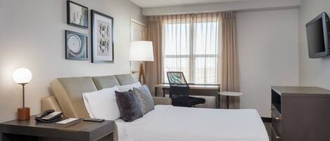 A hotel room with a bed, desk, chair, lamp, TV, and artwork on the wall.