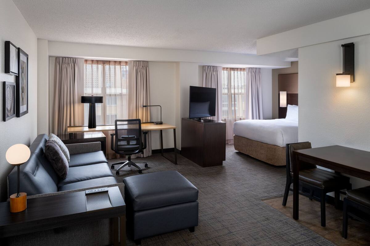 A hotel room with a bed, a desk with a chair, a sofa, an ottoman, a TV, and a lamp.