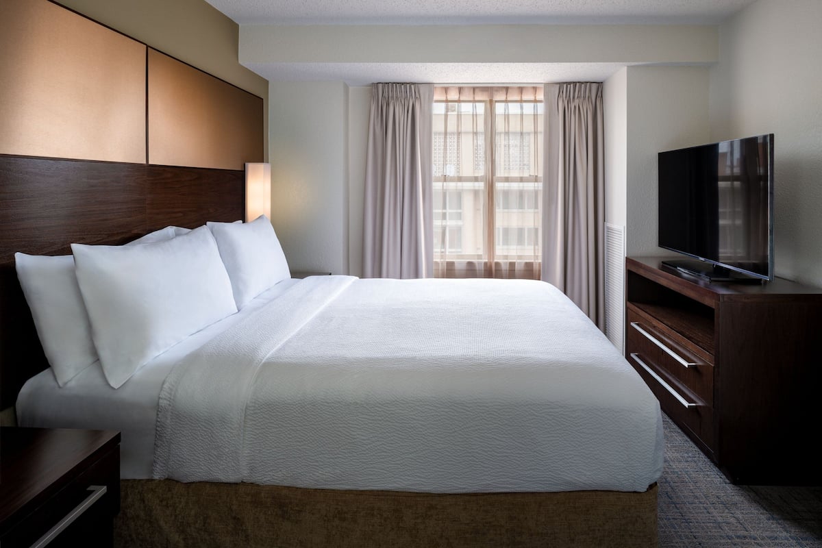 A hotel room with a large bed, a flat-screen TV, a wooden headboard, and a window with curtains.
