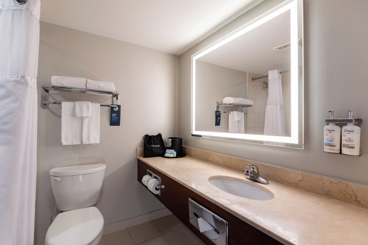 Un baño moderno con inodoro, lavabo y un espejo grande. Hay toallas en un perchero, una cafetera y artículos de aseo en la encimera.