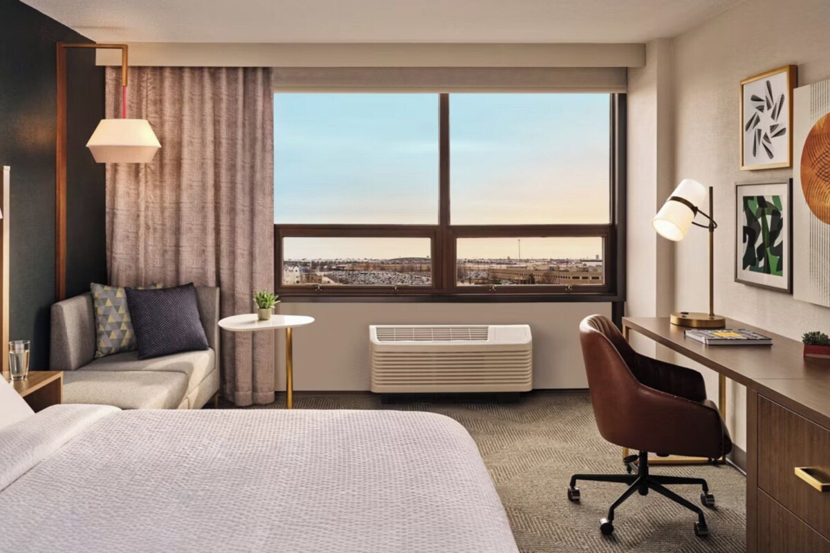 Una habitación de hotel con cama, escritorio, silla y un ventanal con vistas a la ciudad.