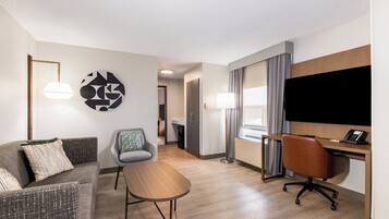 Quarto de hotel moderno com sofá, poltrona, escrivaninha e televisão de tela plana.